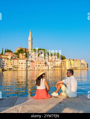 Ein vielseitiges Paar sitzt auf einem Steinpier und bestaunt die malerischen Hügel von Rovinj Kroatien. Das warme Leuchten des Sonnenuntergangs färbt den Himmel, reflektiert vom ruhigen Wasser und schafft einen Moment der Ruhe. Stockfoto