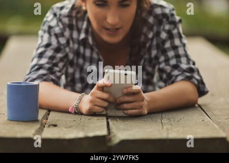 Junge Frau, die auf einem Picknicktisch sitzt, ihr Handy benutzt, während ein Becher neben ihr sitzt, und in der warmen Sommersonne Freizeit-Momente im Freien genießt Stockfoto