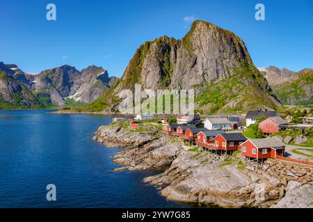 Rote Hütten an der felsigen Küste eines Fjords in Norwegen, umgeben von hohen Bergen und einem klaren blauen Himmel. Hamnoy Fischerdorf auf Lofoten I Stockfoto