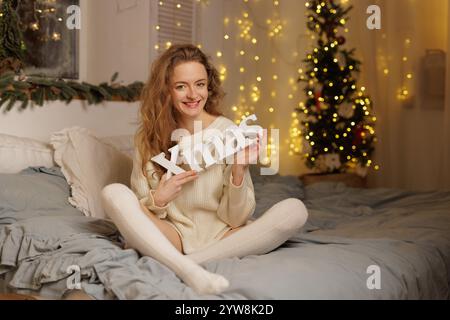 Frau in weißem Strickpullover und Kniestrümpfen mit WEIHNACHTSSCHILD, sitzend auf dem Bett zu Hause am Weihnachtsabend, Zimmer mit Girlanden dekoriert, gemütliches Christm Stockfoto