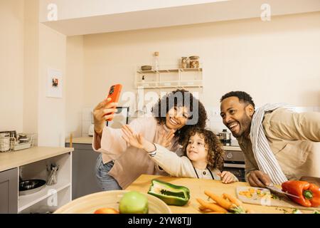 Eine fröhliche Familie während eines Videoanrufs in der Küche. Die Eltern und das Kind lächeln und genießen ihre gemeinsame Zeit, schauen auf das Smartphone und Stockfoto