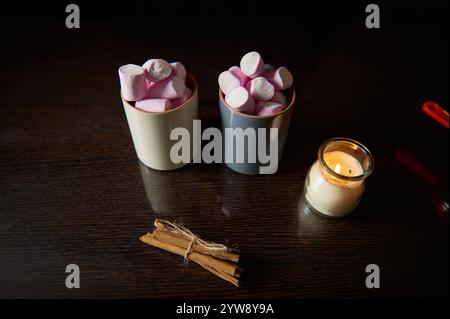 Zwei Tassen gefüllt mit rosa Marshmallows neben einer beleuchteten Kerze und Zimtstangen schaffen eine warme, gemütliche Atmosphäre. Ideal für saisonale Einrichtung, Entspannungskoje Stockfoto