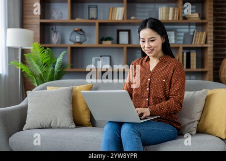 Asiatische Frau, die zu Hause auf dem Laptop arbeitet und bequem auf dem Sofa sitzt. Freizeitkleidung sorgt für eine entspannte Atmosphäre. Konzentriert sich auf Aufgaben in gemütlicher Wohnzimmeratmosphäre. Ideal für Home-Office-Themen. Stockfoto