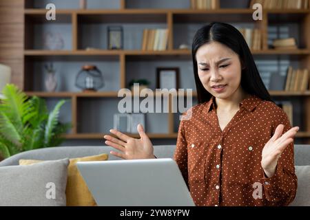 Die asiatische Frau zu Hause sieht frustriert aus, wenn sie an einem Laptop arbeitet. Sie drückt negative Emotionen mit erhobenen Händen aus, während sie im modernen Wohnzimmer sitzt. Frustrationskonzept und Technologiefragen. Stockfoto