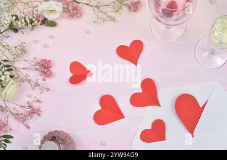 Rote Herzen fallen aus dem Umschlag, Komposition umgeben von Rosen, Pfingstrosen, gypsophila-Blüte und Weingläsern auf rosa Hintergrund. Stockfoto