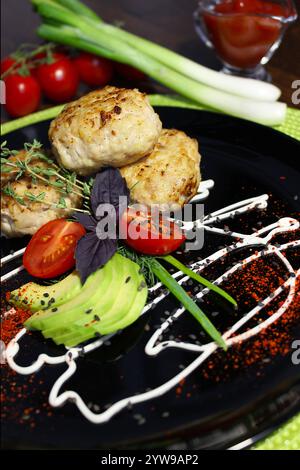 Hähnchenkoteletts auf einem schwarzen Teller mit Avocado, Tomaten und einer kreativen Soße Stockfoto