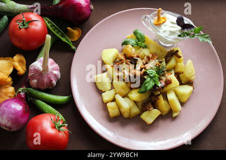 Gebratene Kartoffelscheiben mit Pfifferlingen, Zwiebeln, Dill, Basilikum und Sauerrahm werden auf einem rosafarbenen Teller serviert, umgeben von frischem Gemüse Stockfoto