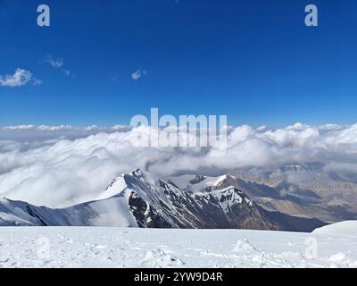 Schneebedeckte Berggipfel erheben sich dramatisch gegen einen leuchtend blauen Himmel mit Wolken, die sich am Horizont ausbreiten. Stockfoto