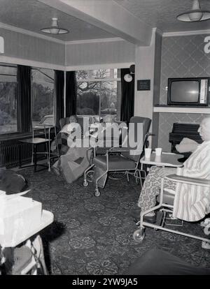 1970er Jahre, historische, ältere Patienten sitzen in medizinischen Stühlen mit hohem Rücken und Teppichen darüber in einer Lounge oder einem Ruheraum eines Altenheims oder Pflegeheims, England Großbritannien. Klavier in der Ecke. Tragbarer Fernseher der Ära in einem Regal darüber. Stockfoto