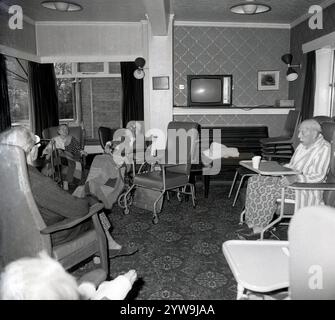 1970er Jahre, historische, ältere Patienten sitzen in medizinischen Stühlen mit hohem Rücken und Teppichen darüber in einer Lounge oder einem Ruheraum eines Altenheims oder Pflegeheims, England Großbritannien. Zur Unterhaltung stehen ein Klavier in der Ecke und ein tragbarer Fernseher der Epoche auf einem Regal darüber. Stockfoto