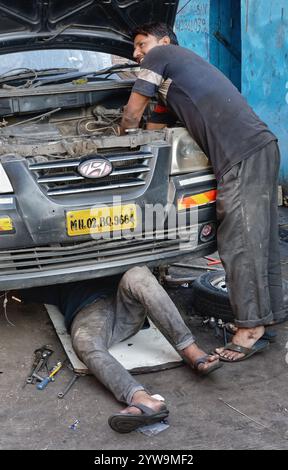 Zwei junge indische Autoreparaturmänner in Mumbai, Indien, arbeiten am Motor eines Autos, einer von ihnen arbeitet unter dem Fahrzeug Stockfoto