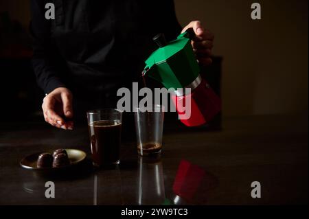 Eine Person gießt frisch gebrühten Kaffee aus einer lebendigen Mokakanne in Gläser, begleitet von Schokoladengerichten an der Seite. Stockfoto