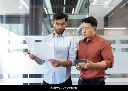 Zwei männliche Fachleute diskutieren über Papierkram in einer modernen Büroumgebung. Sie wirken fokussiert und engagiert und betonen Teamarbeit und Kommunikation in einem Unternehmensumfeld. Stockfoto