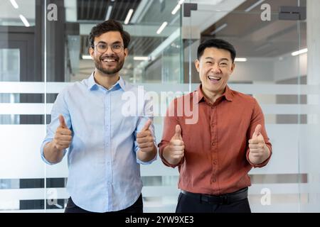 Zwei lächelnde Männer in einem Büro, die mit gedrückten Daumen positiv zum Ausdruck kommen und Erfolg und Teamarbeit symbolisieren. Das Bild vermittelt eine freundliche und professionelle Atmosphäre Stockfoto