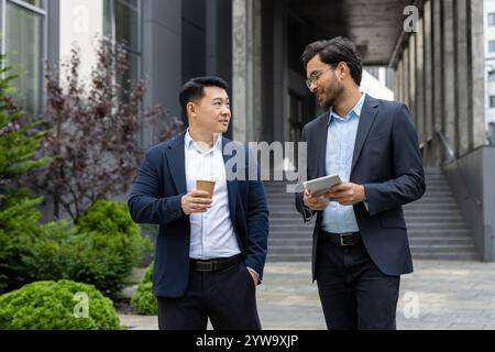 Zwei professionelle Männer in Geschäftskleidung führen ein freundliches Gespräch, während sie Kaffeetassen und Tablets halten. Die Umgebung ist städtisch, mit Grün und moderner Architektur im Hintergrund. Stockfoto