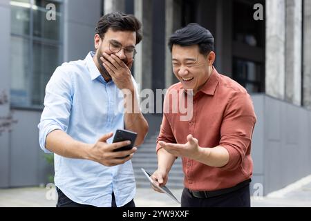 Zwei Männer teilen einen freudigen Moment, lachen, während sie etwas auf einem Smartphone anschauen. Sie scheinen vor einem Bürogebäude zu sein und genießen eine Pause miteinander. Stockfoto