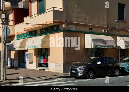 Savona. Italia - 10. Dezember 2024: Apotheke an einer sonnigen Straßenecke in Savona, Italien, mit geparkten Autos und einer Fußgängerüberfahrt davor Stockfoto