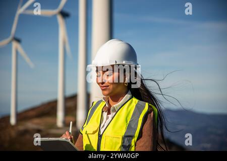 latein weibliche Ingenieurin mit Sicherheitsweste und Helm macht Notizen auf einem Tablet, während sie Windräder inspiziert und so zur Entwicklung sauberer Energie beiträgt Stockfoto