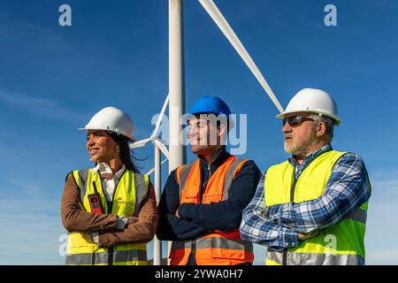 Drei multiethnische Ingenieure, die Sicherheitswesten und Helme tragen, stehen mit überkreuzten Armen in einem Windkraftwerk, das erneuerbare ener repräsentiert Stockfoto