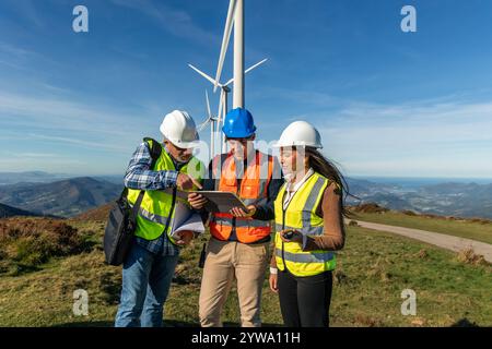 Drei multiethnische Ingenieure, die Sicherheitswesten und Helme tragen, verwenden ein digitales Tablet und diskutieren neben Windturbinen auf einem Hügel mit Blick Stockfoto