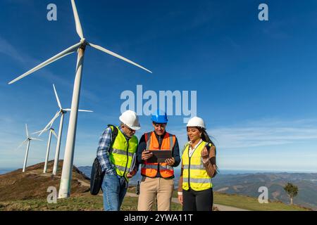 Drei multiethnische Ingenieure, die Sicherheitswesten und Helme tragen, verwenden ein Tablet, diskutieren und planen neben Windturbinen auf einem Hügel und arbeiten Stockfoto