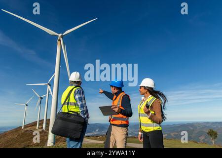 Drei multiethnische Ingenieure, die Sicherheitswesten und Helme tragen, diskutieren neben einer Windkraftanlage, was zu sauberer Energie und Umwelteinsparungen beiträgt Stockfoto