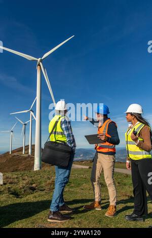 Drei multiethnische Ingenieure, die Sicherheitswesten und Helme tragen, diskutieren in der Nähe von Windturbinen und arbeiten gemeinsam an einem Projekt, das sich auf grüne Energie konzentriert. Stockfoto