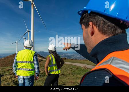 Drei multiethnische Ingenieure, die Sicherheitswesten und Helme tragen, stehen auf einem Berggipfel in der Nähe von Windturbinen, wobei ein Techniker auf den zeigt Stockfoto