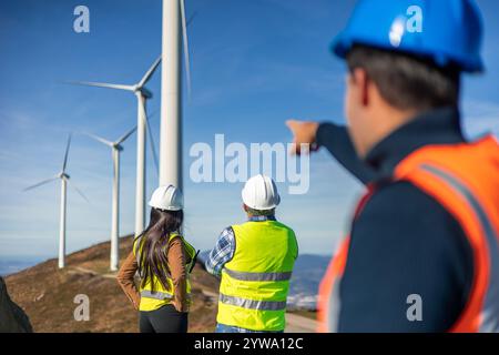 Drei multiethnische Ingenieure, die Sicherheitswesten und Helme tragen, zeigen auf Windräder auf einem Hügel und diskutieren über die Entwicklung erneuerbarer Energien Stockfoto