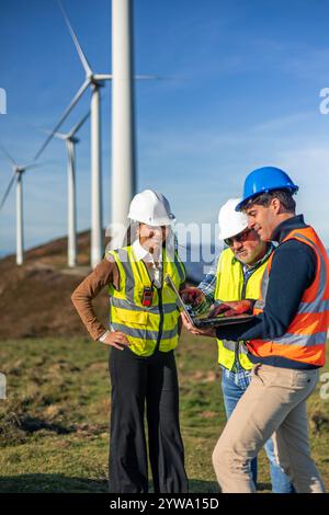 Drei multiethnische Ingenieure, die Sicherheitswesten und Helme tragen, benutzen einen Laptop und arbeiten an einem Projekt in einem Windkraftwerk zusammen Stockfoto