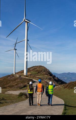 Drei multiethnische Ingenieure, die Sicherheitswesten und Helme tragen, gehen auf einer asphaltierten Straße auf einem Berggipfel, überprüfen den Zustand von Windrädern, Stockfoto