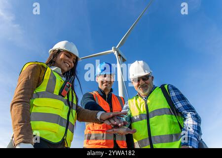 Drei multiethnische Ingenieure, die Sicherheitswesten und Helme tragen, schließen sich vor einer Windkraftanlage zusammen und symbolisieren Teamarbeit und Engagement für rene Stockfoto