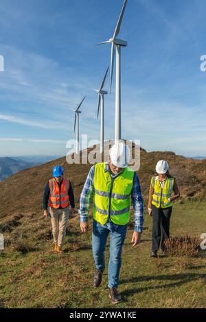 Drei multiethnische Ingenieure, die Sicherheitswesten und Helme tragen, gehen bergab in einem Windkraftwerk auf einem Berggipfel und inspizieren den Betrieb Stockfoto