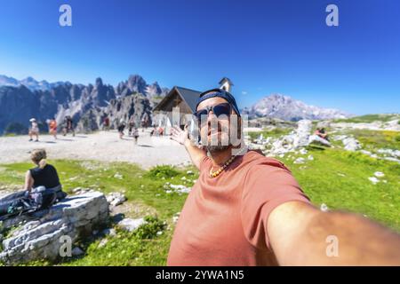 Touristen machen ein Selfie mit offenen Armen vor der Kapelle auf dem Wanderweg Tre cime di lavaredo in den italienischen dolomiten. Cappella degli Alpini Stockfoto