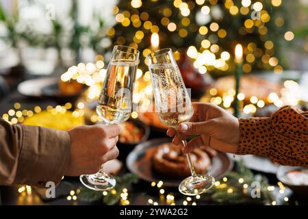 Zwei Personen zwitschern zur Feier Sektgläser, mit einem festlichen Tisch für ein Festtagsessen im Hintergrund, geschmückt mit Lichtern und Dekorationen Stockfoto