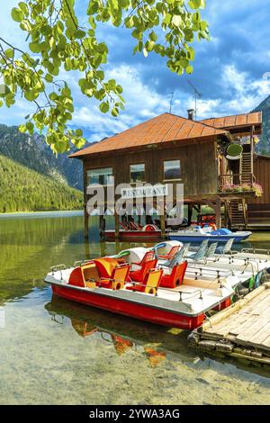 Farbenfrohe Tretboote liegen unter einem Holzrestaurant am toblacher See an, an einem klaren Sommertag erheben sich die dolomiten im Hintergrund Stockfoto