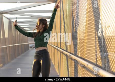 Glückliche junge Frau tanzt und filmt sich mit ihrem Handy auf der Straße bei Sonnenuntergang. Stockfoto