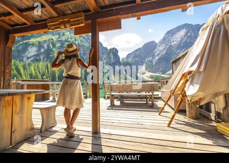 Frau mit Strohhut, die auf einem Holzsteg steht und die atemberaubende Landschaft des pragser Sees und der umliegenden Berge in den dolomiten bewundert Stockfoto