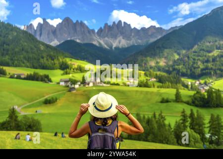 Weibliche Touristenin, die ihren Hut hält, während sie auf die Straße blickt. Die Magdalena-Kirche im val di funes mit den Bergen im Hintergrund Stockfoto