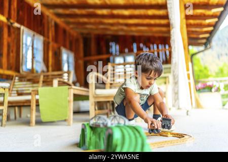 Kind, das sich mit Spielzeugautos auf einer sonnigen Terrasse im Freien, umgeben von Holzmöbeln, einlässt Stockfoto