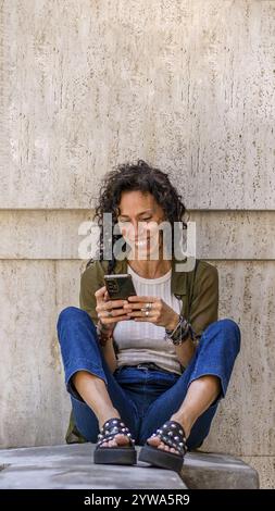 Frau im mittleren Alter, die auf einem Felsvorsprung sitzt, während sie ihr Smartphone benutzt Stockfoto