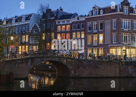 Abendlicher Blick auf die Stadt mit beleuchtetem Kanal und Brücke voller Fahrräder, Amsterdam, Niederlande Stockfoto