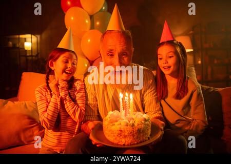 Fröhliche Familiengeburtstagsfeier mit Großvater und Enkelinnen, die einen besonderen Kuchen-Moment im Haus teilen Stockfoto