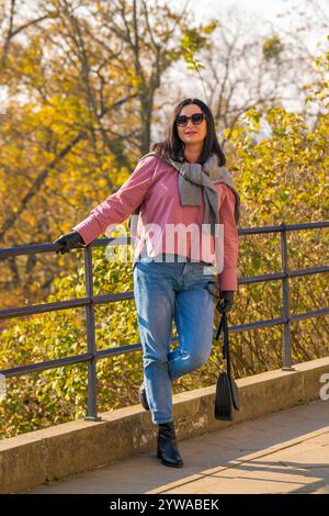 Hübsche Frau mit dunklem Haar, rosa Pullover, Jeans, Sonnenbrille, schöne Herbstlandschaft im Park. Kaukasische Frau schaut nachdenklich in di Stockfoto