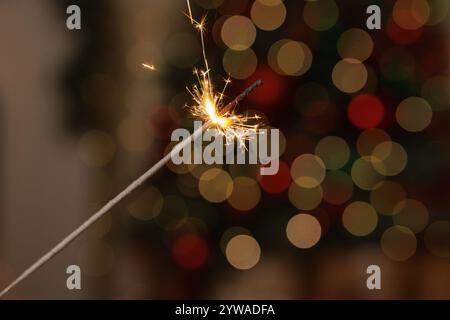 Brennender Funkler in weiblicher Hand auf dem Hintergrund von weihnachtsbaumlichtern im dunklen Raum. Frohes Neues Jahr! Hand hält Feuerwerk vor stilvoll dekoriertem T Stockfoto