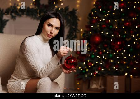 Eine Frau in einem kuscheligen weißen Pullover hält ein rotes Weihnachtsschmuck, während sie in einer festlichen Umgebung mit einem geschmückten Weihnachtsbaum im Hintergrund sitzt Stockfoto