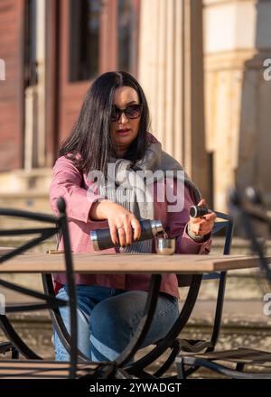 Hübsche Frau in Sonnenbrille sitzt am Tisch in einem Café im Freien und gießt sich Tee oder Kaffee aus der Thermoskanne und genießt den letzten sonnigen Herbsttag, freien Tag Stockfoto