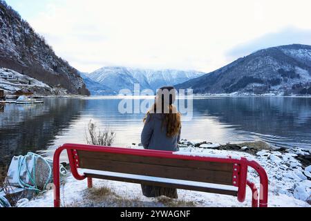 Eine Frau mit Hut und Mantel sitzt auf einer verschneiten Bank und bewundert die beschauliche Fjord- und Berglandschaft in Gaupne, Norwegen Stockfoto