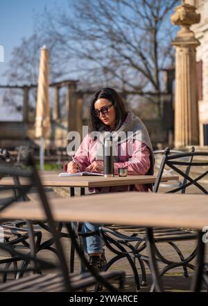 Hübsche Frau, die in einem Café im Freien sitzt und Notizen in Notizbuch macht. In der Nähe auf dem Tisch befindet sich eine Thermoskanne mit brennendem Getränk. Brünette arbeitet ferngesteuert, denk Stockfoto