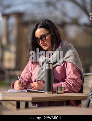 Hübsche Frau, die in einem Café im Freien sitzt und Notizen in Notizbuch macht. In der Nähe auf dem Tisch befindet sich eine Thermoskanne mit brennendem Getränk. Brünette arbeitet ferngesteuert, denk Stockfoto